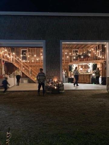 People dining and socializing inside a warmly lit barn-style venue with a staircase, visible through large open doors at night.