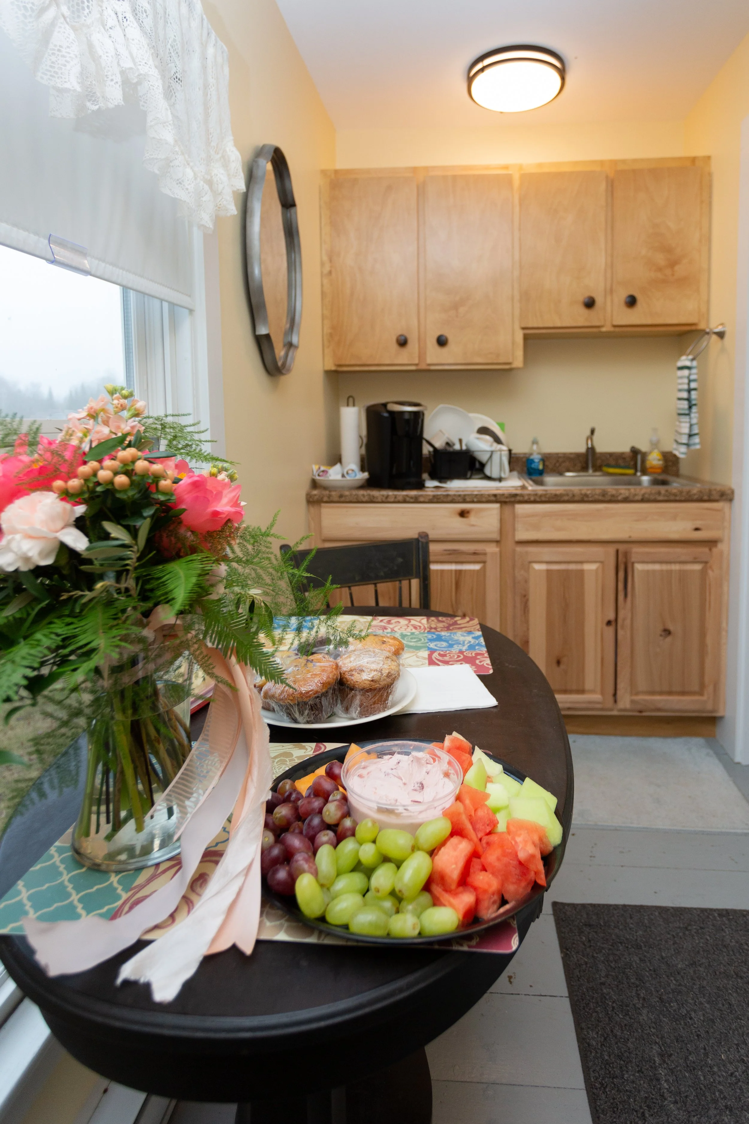 A dining table with a bouquet of pink flowers and a plate of muffins, surrounded by cut watermelon, cantaloupe, grapes, and a bowl of dip, in a kitchen with wooden cabinets and a coffee maker.