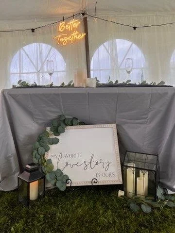 Wedding decor with a white table draped with a white cloth, surrounded by lanterns with candles, a framed sign reading 'my favorite love story is ours' adorned with eucalyptus, and a neon sign that says 'Better Together' hanging above.