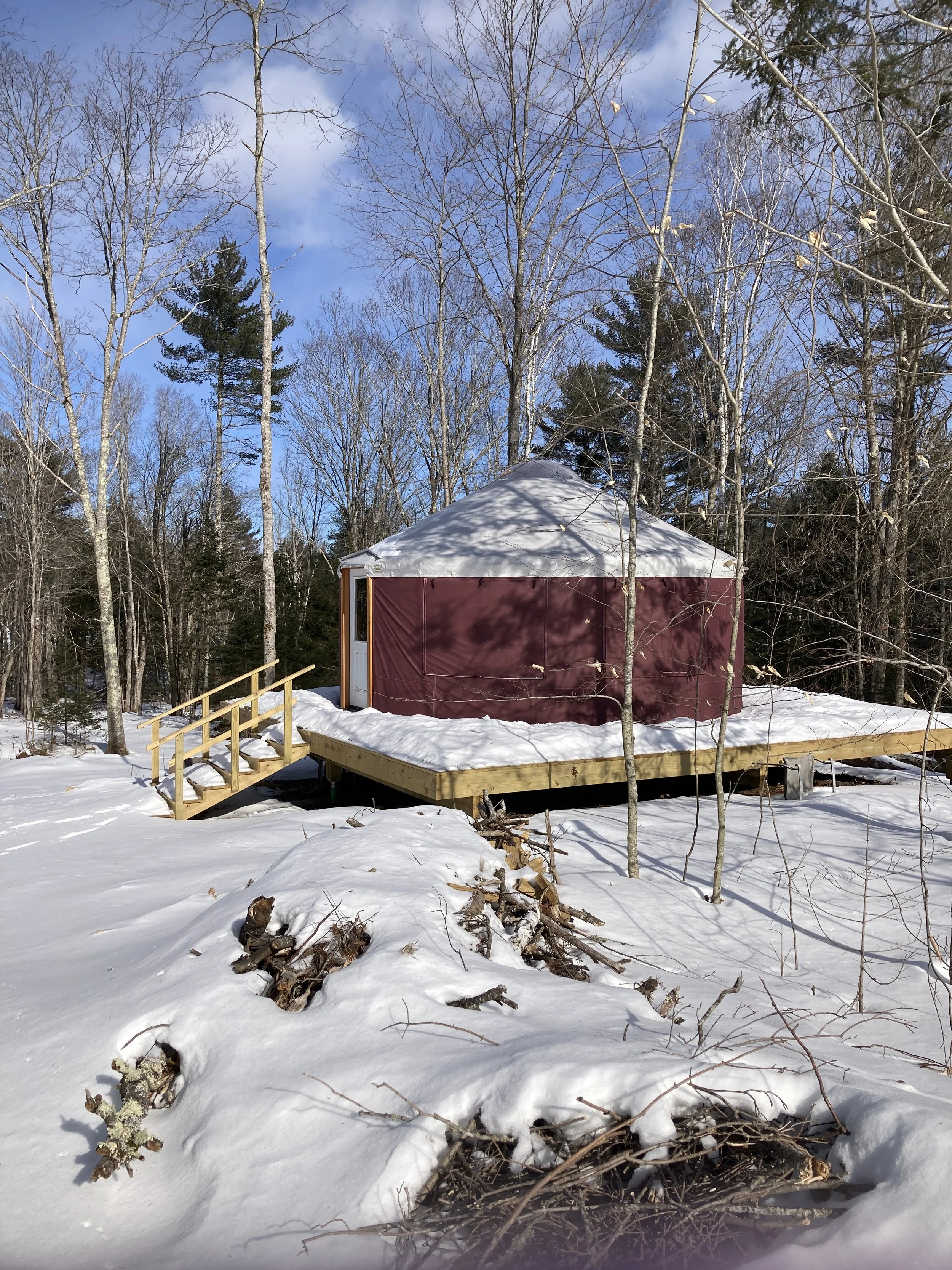 A small round tent with a white top and maroon walls is built on a wooden deck in a snowy forest. The deck has a small set of stairs leading up to it.