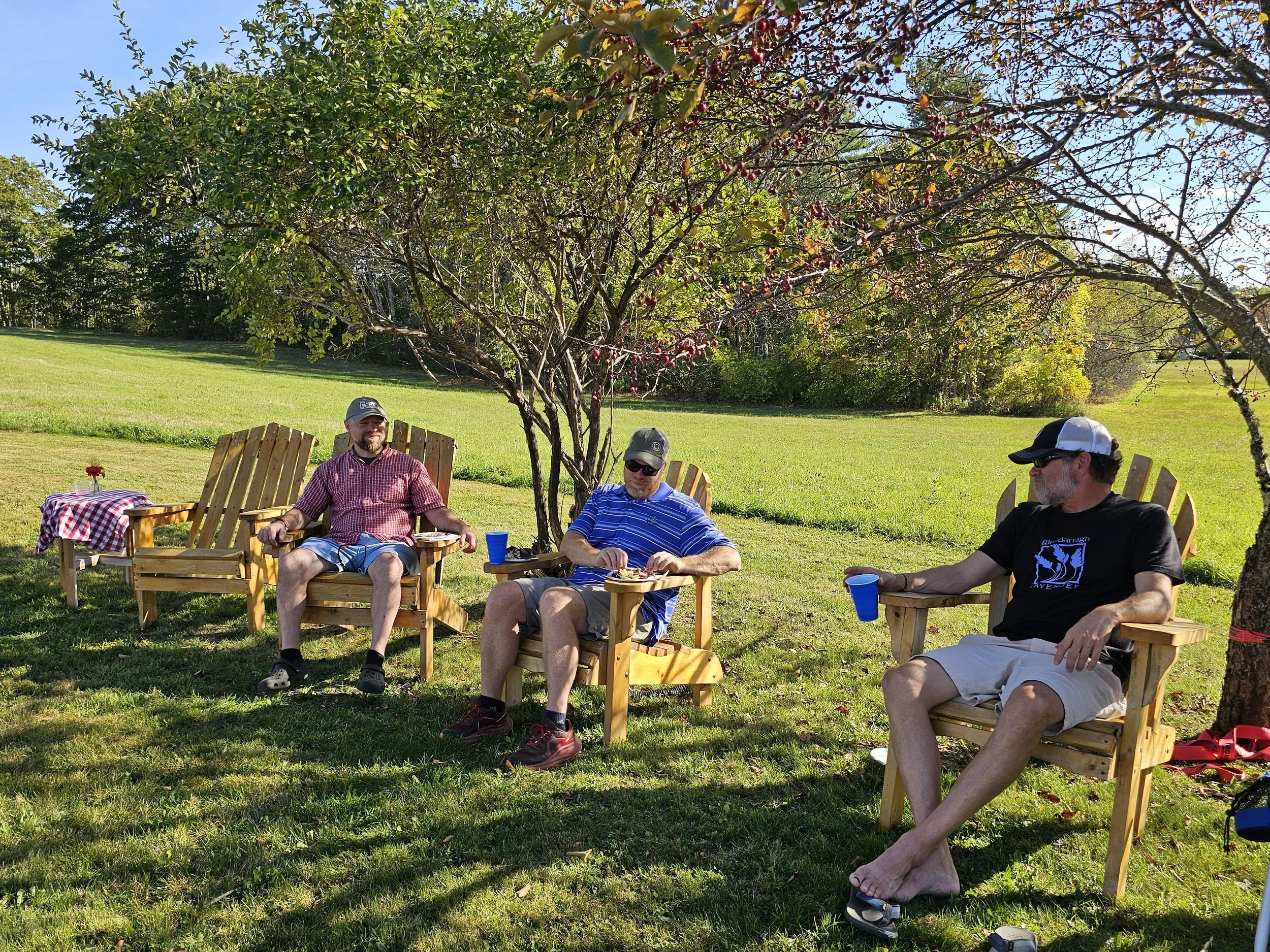 Four men sitting outdoors in wooden chairs under a tree on a sunny day, enjoying food and drinks, with a grassy field and trees in the background.