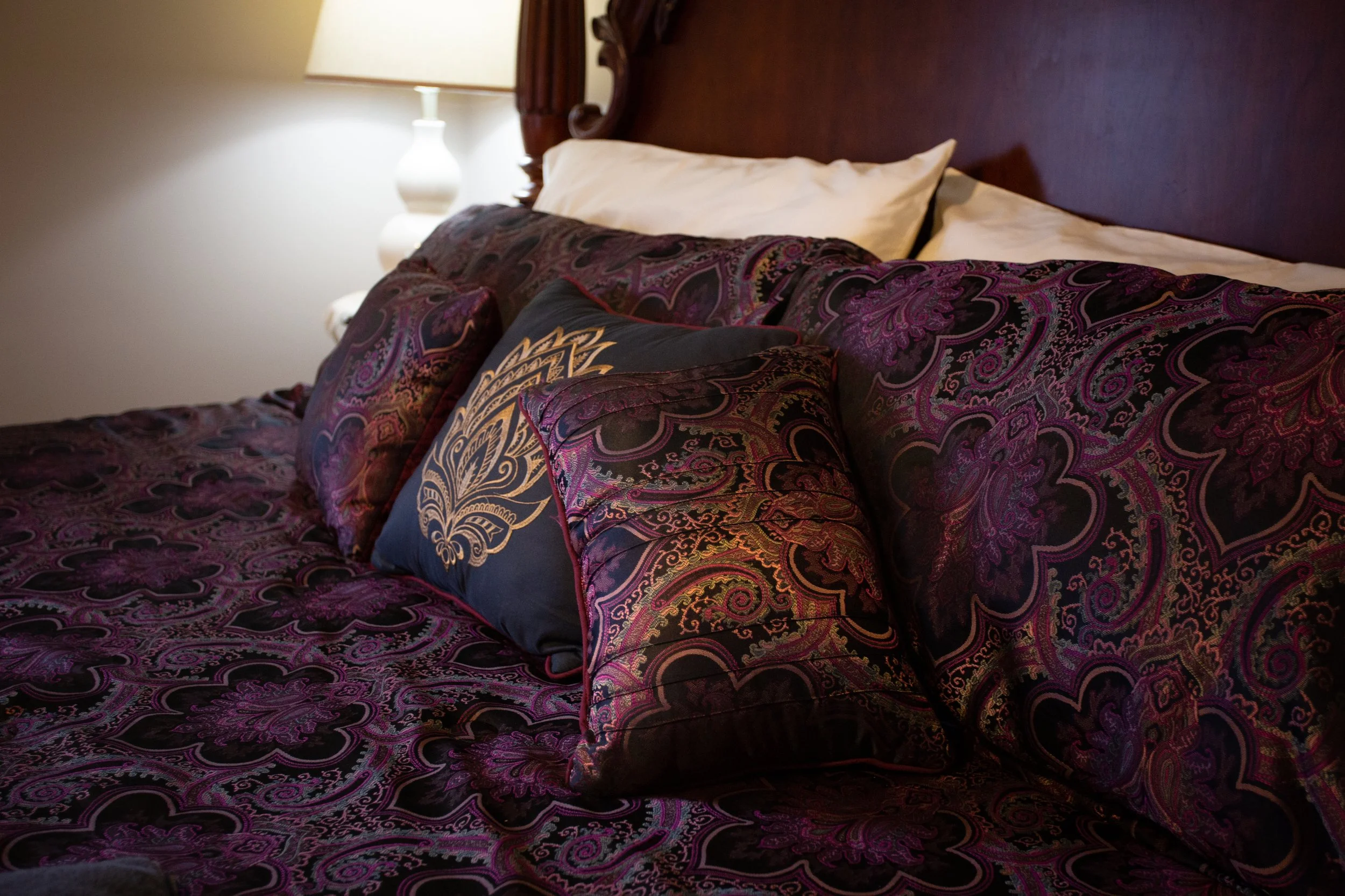 A bed with dark purple and black paisley-patterned bedding, including multiple pillows and a headboard. A nightstand with a lamp is partially visible on the side.