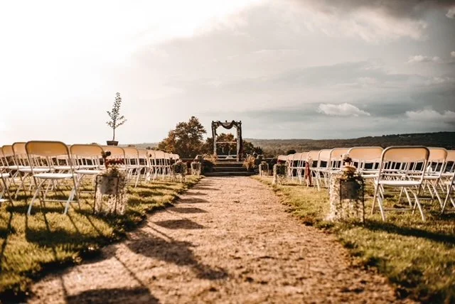 Outdoor wedding ceremony setup with rows of chairs on either side of a dirt aisle and a wooden arch at the front under cloudy sky.