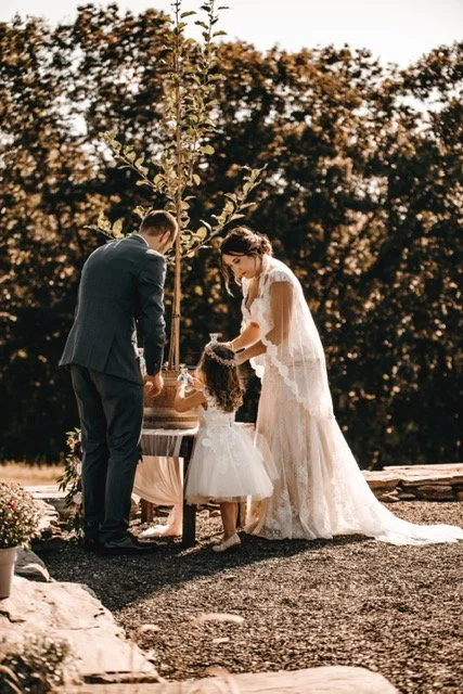 A couple, a man and a woman, and a young girl standing outdoors in front of a small table, with trees in the background, during a special event like a wedding.