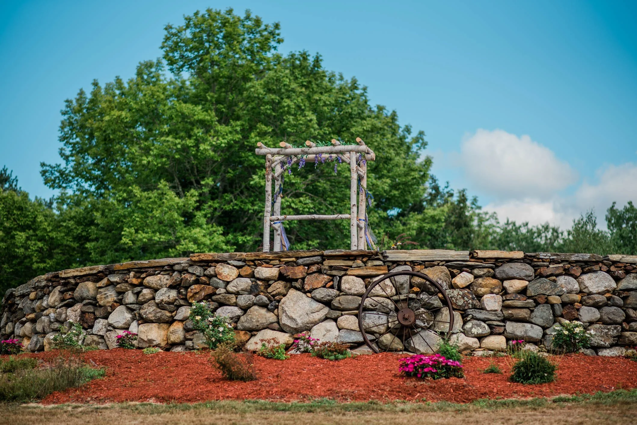 A stone wall with a rustic wooden structure on top, vintage wagon wheel leaning against the wall, and flowers in front, set against green trees and a blue sky with clouds.