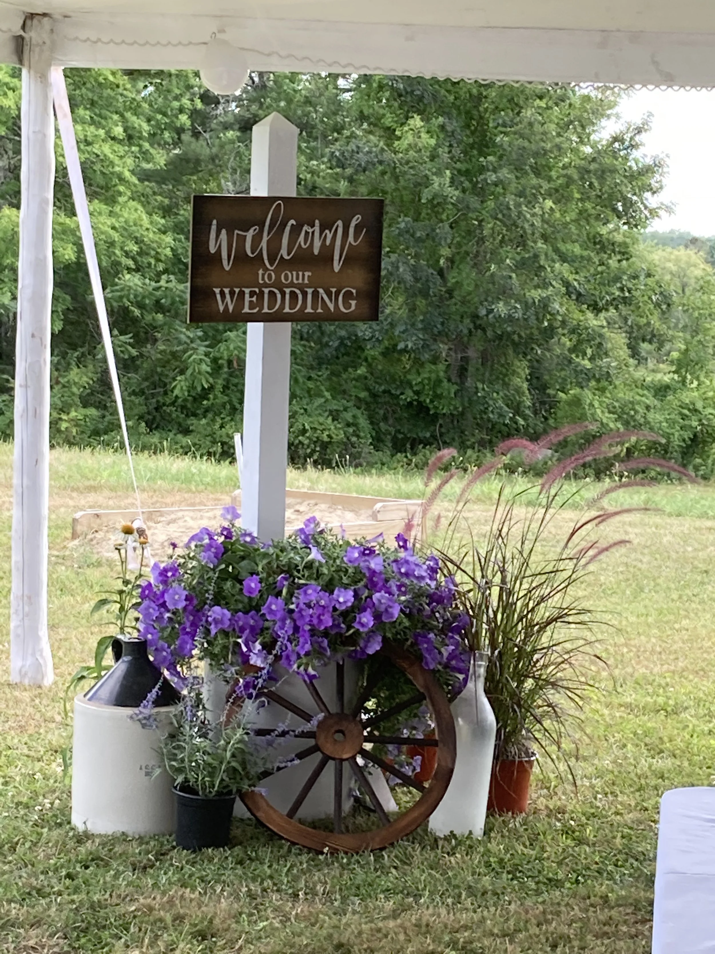 Wedding welcome sign with purple flowers, an old wagon wheel, and decorative vases on the grass.