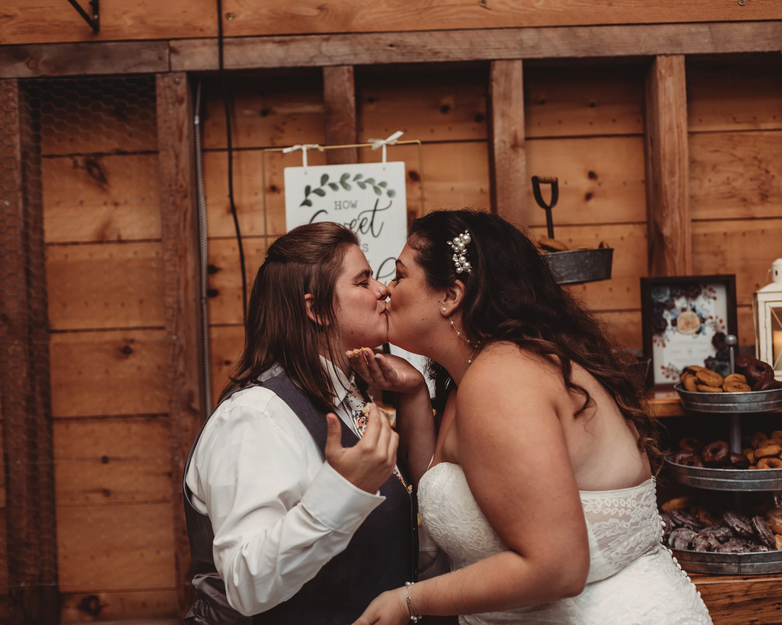 Two women sharing a kiss at a wedding reception, one in a white wedding dress and the other in formal attire, with a wooden backdrop and bakery items in the background.
