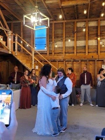 Couple dancing at a wedding reception in a rustic barn with wooden walls and warm lighting, surrounded by guests watching and taking photos.
