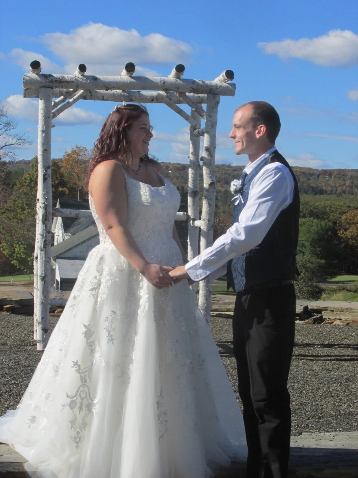 A bride and groom hold hands and smile at each other during their wedding ceremony outdoors, with a blue sky and scenic landscape in the background.