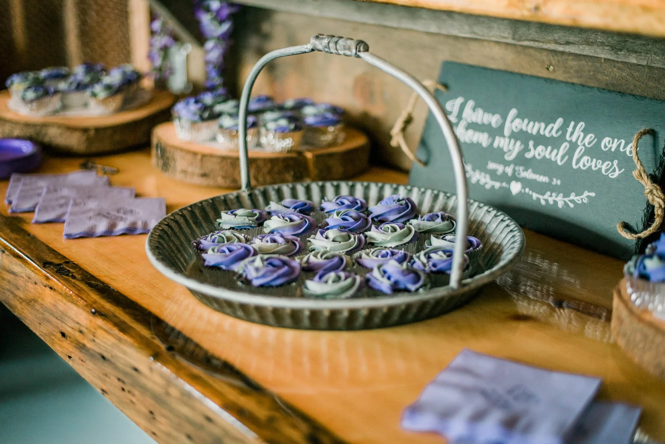 A rustic wooden table displays a metal basket filled with purple and green fabric roses, with lavender-colored handkerchiefs beside it. In the background, there are wooden log slices holding small containers of matching fabric roses. A decorative sig