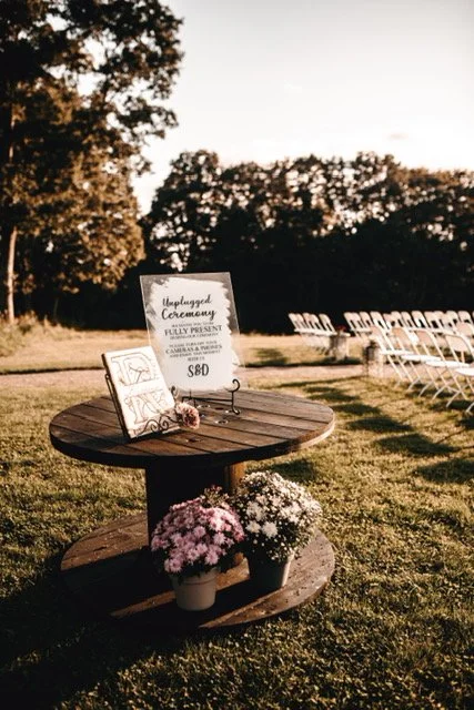Outdoor wedding setup with a round wooden table holding flowers and an unplugged wedding sign, with chairs arranged in the background on a grassy field during sunset.