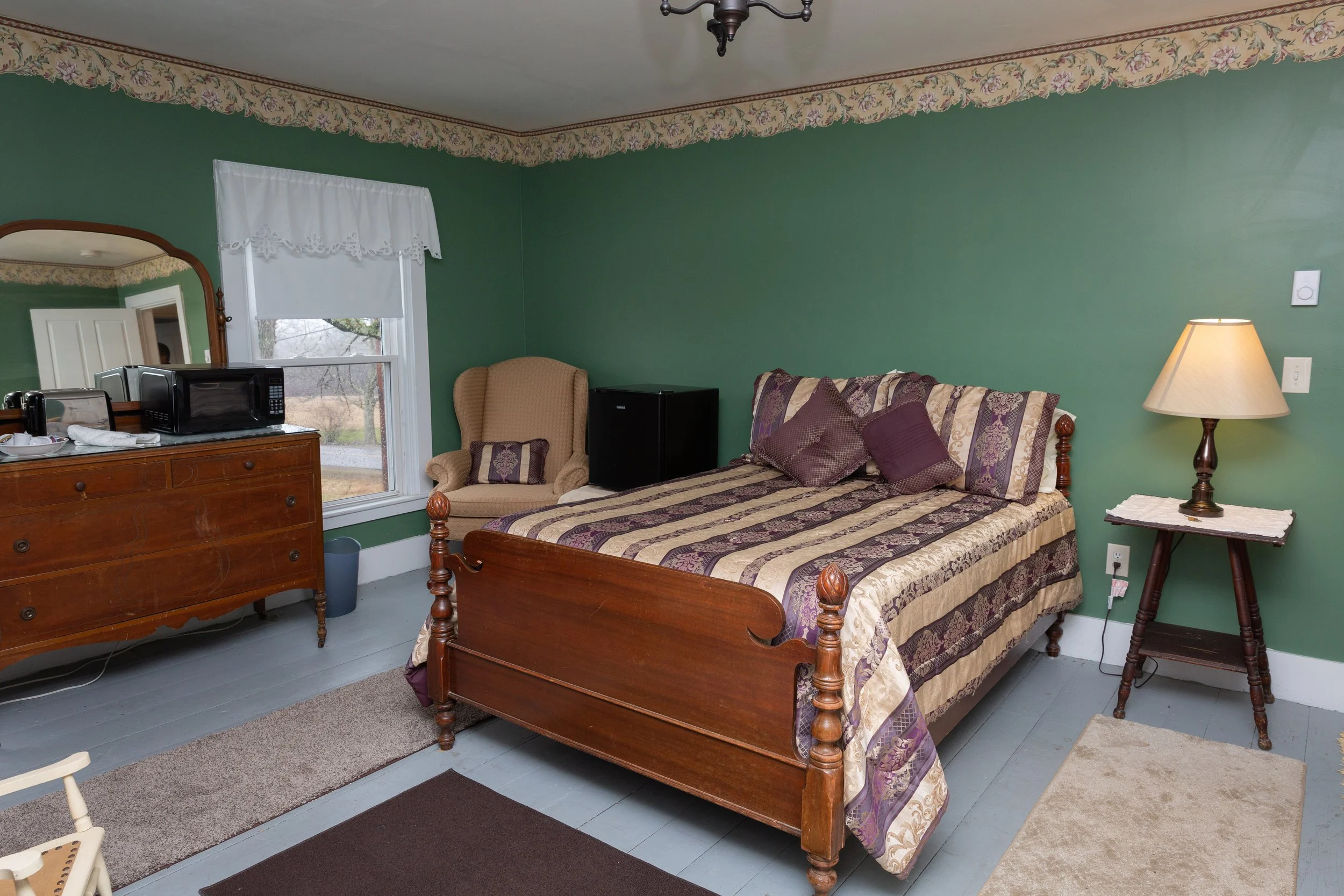 A bedroom with green walls, a wooden bed with floral bedspread, a bedside table with a lamp, an armchair near a window, a dresser with a microwave, a mini refrigerator, and various rugs on light blue flooring.