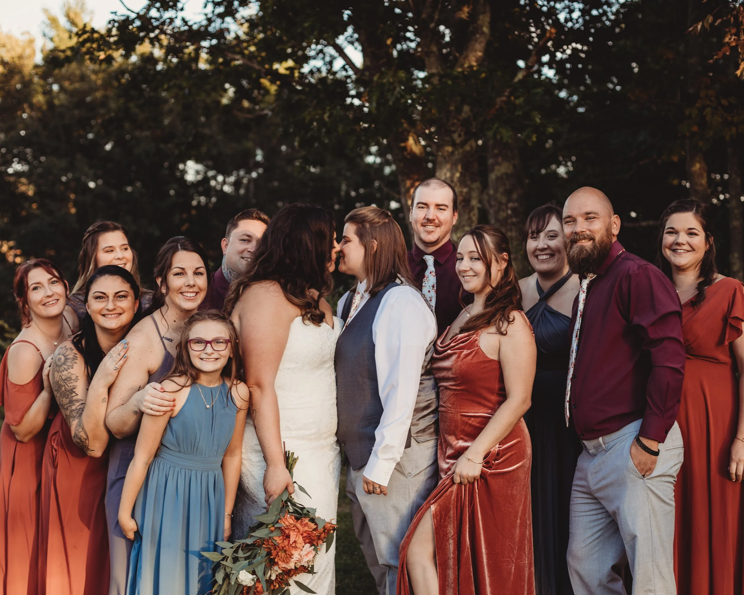 A wedding celebration with a diverse group of people, including a bride and groom, standing outdoors at sunset with trees in the background.