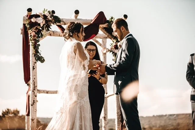 Bride and groom exchanging vows under an outdoor wooden arch decorated with flowers, with officiant standing beside them on a sunny day.