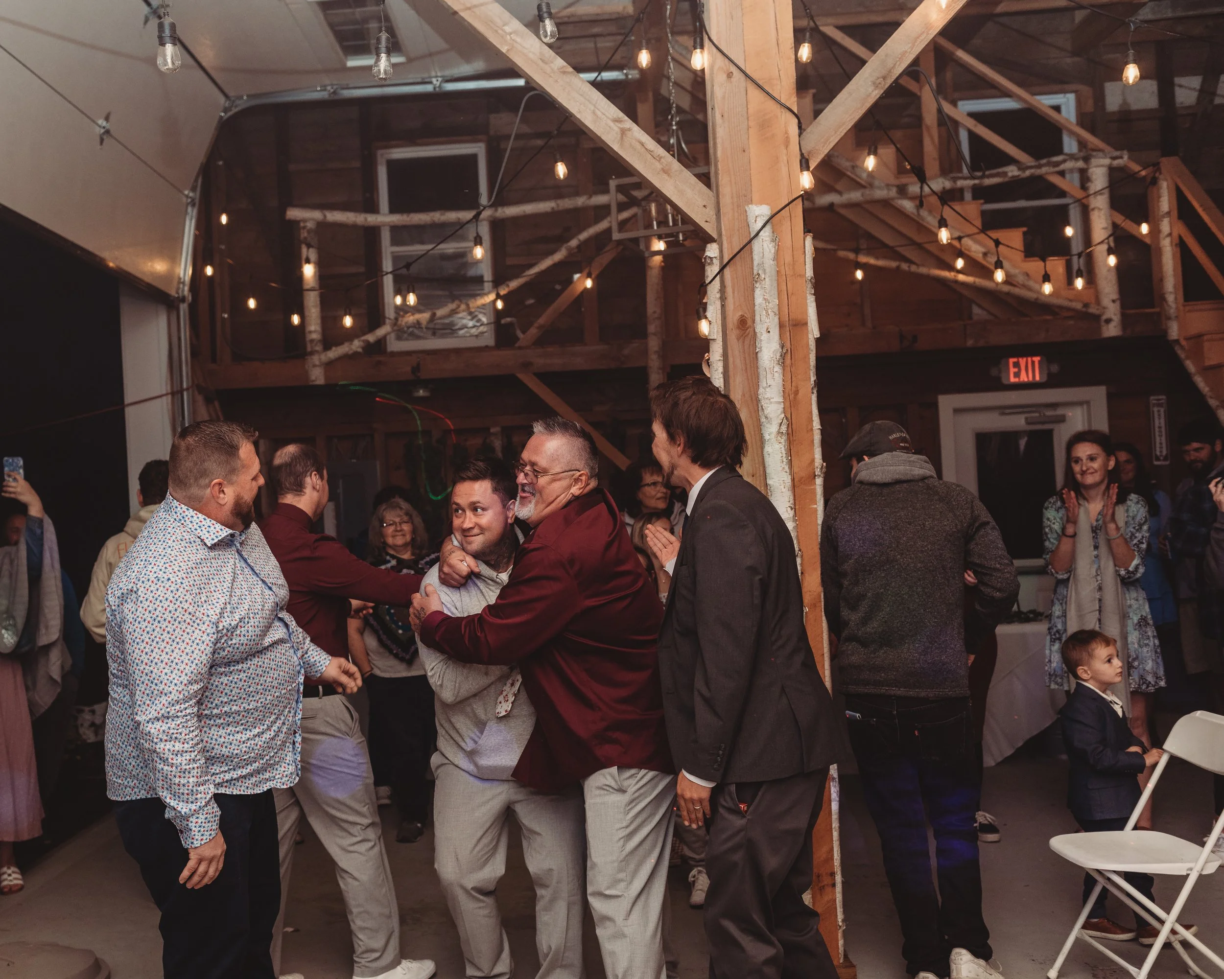 People dancing and celebrating at an indoor event with string lights and wooden beams.