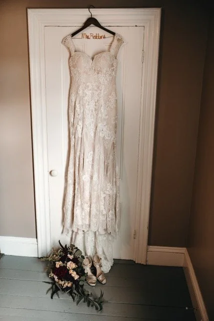 A wedding dress hanging on a hanger with the words 'Mrs. Ratlin' on it, with a bouquet and shoes on the floor beneath.