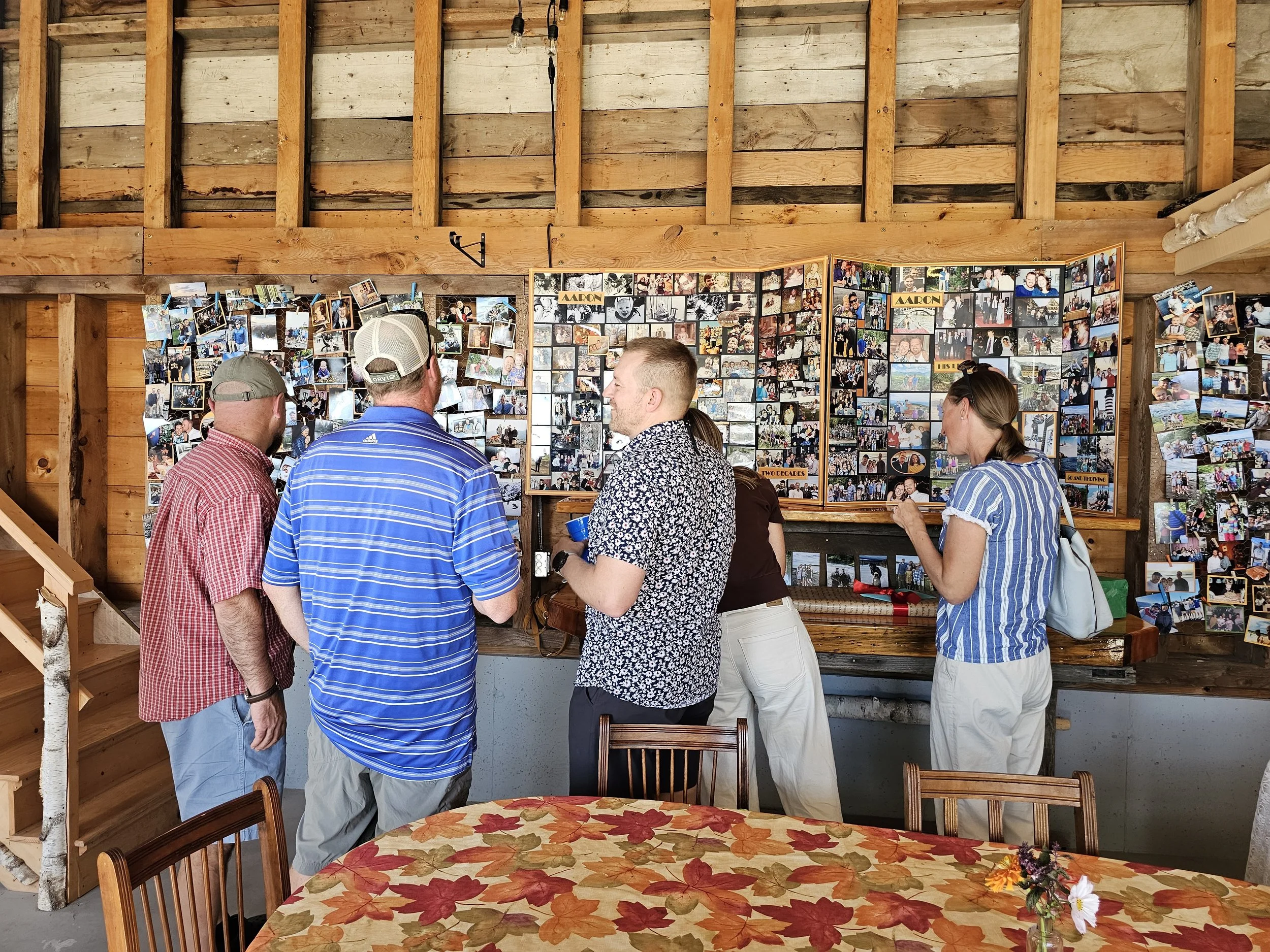 People gather around a display of numerous photographs on a wall in a rustic wooden boathouse or barn. The photos seem to be of various moments, possibly for a celebration or memorial. A table with a fall-colored tablecloth and a small floral arrange