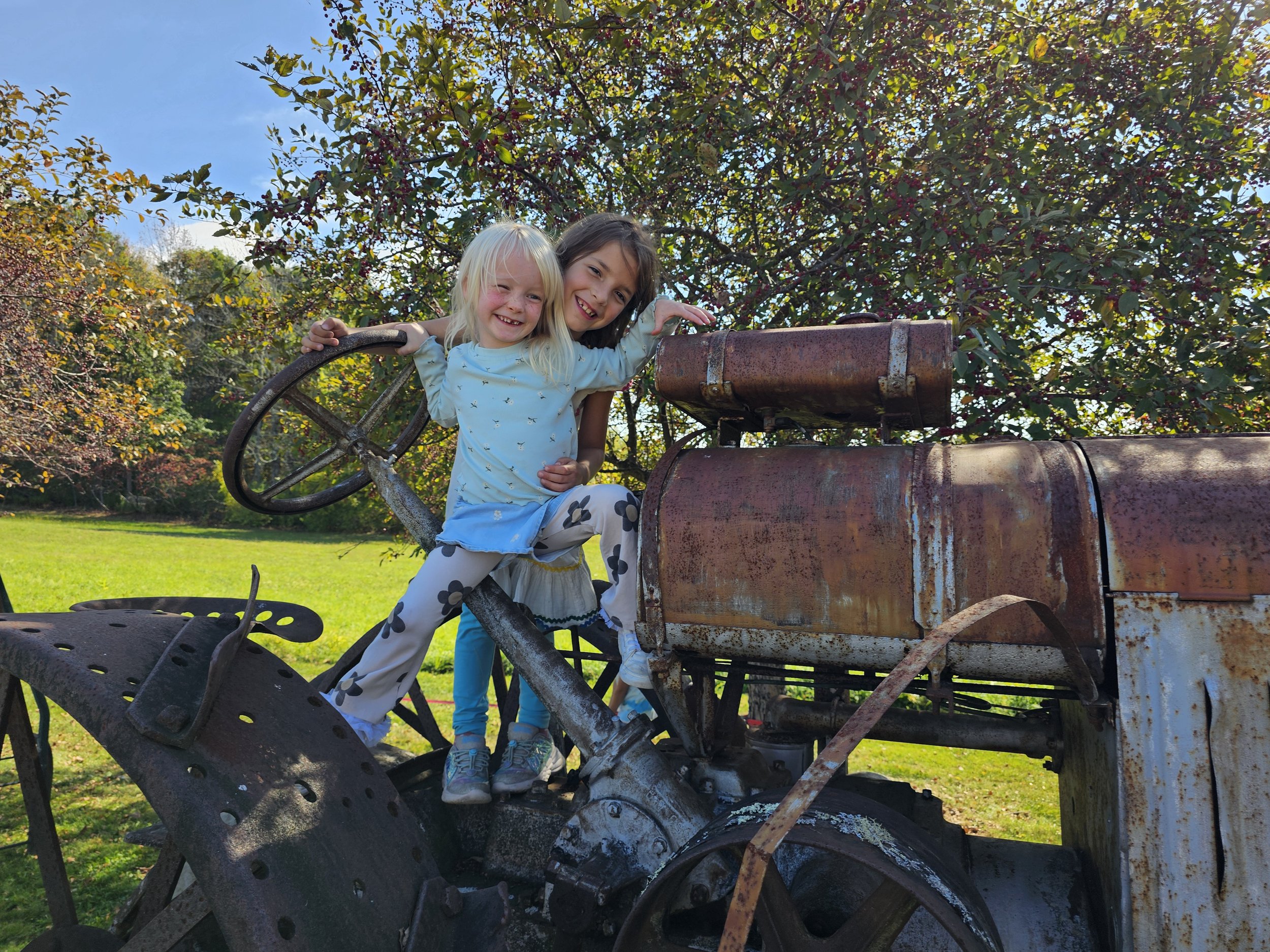 Two young girls sitting on an old rusty tractor outdoors with trees and blue sky in the background. They are smiling and appear happy.