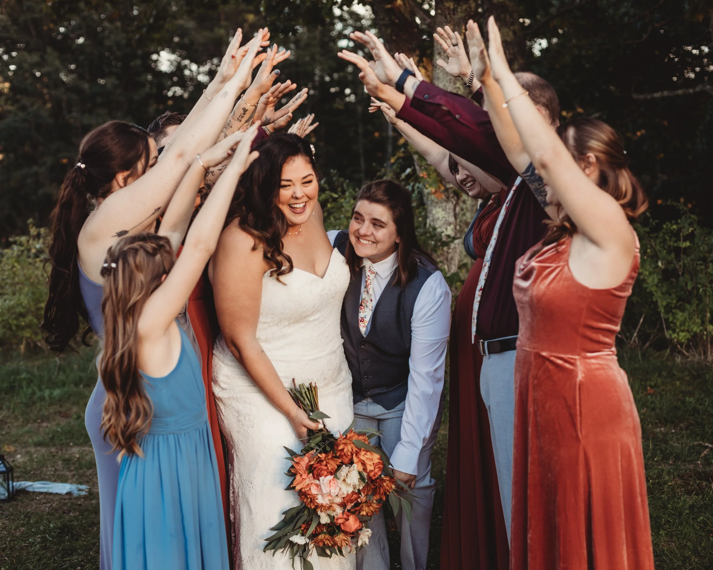 Group of friends celebrating at outdoor wedding, woman in white dress holding bouquet, surrounded by friends raising hands in celebration.