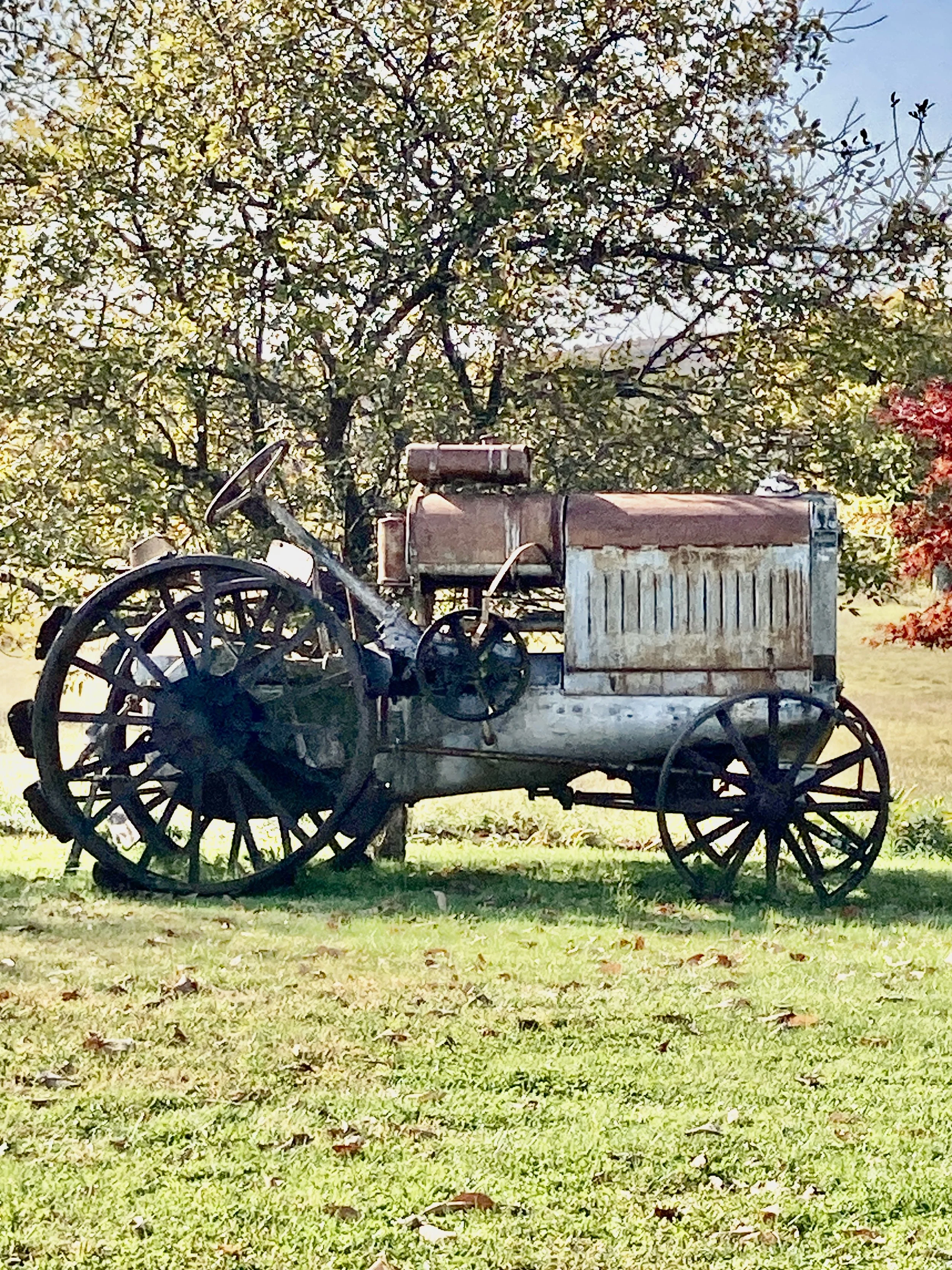 An old rusty tractor with large spoked wheels sitting on a grassy area, with leafy trees and a partly cloudy sky in the background.