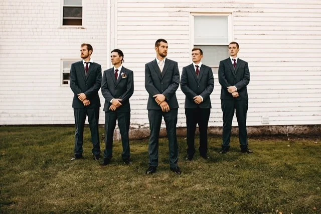 Five men in suits standing outdoors on grass in front of a white wooden building.