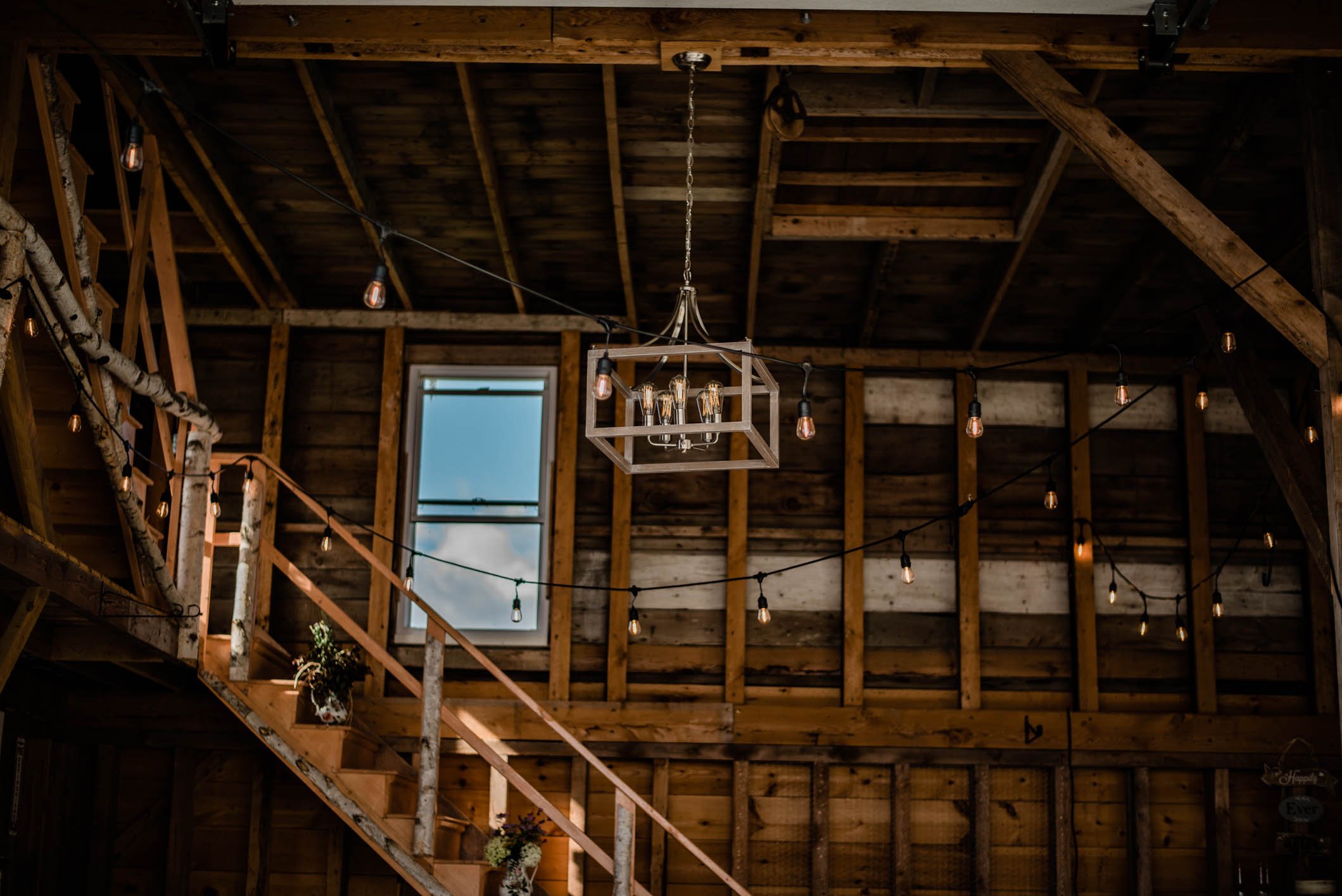 Interior view of a rustic wooden barn with string lights, a window showing sky and clouds, a chandelier, and a staircase with potted plants.