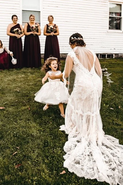 Bride in a lace wedding gown interacts with a young flower girl in a white dress, while three bridesmaids in dark dresses admire in the background outside a white house.