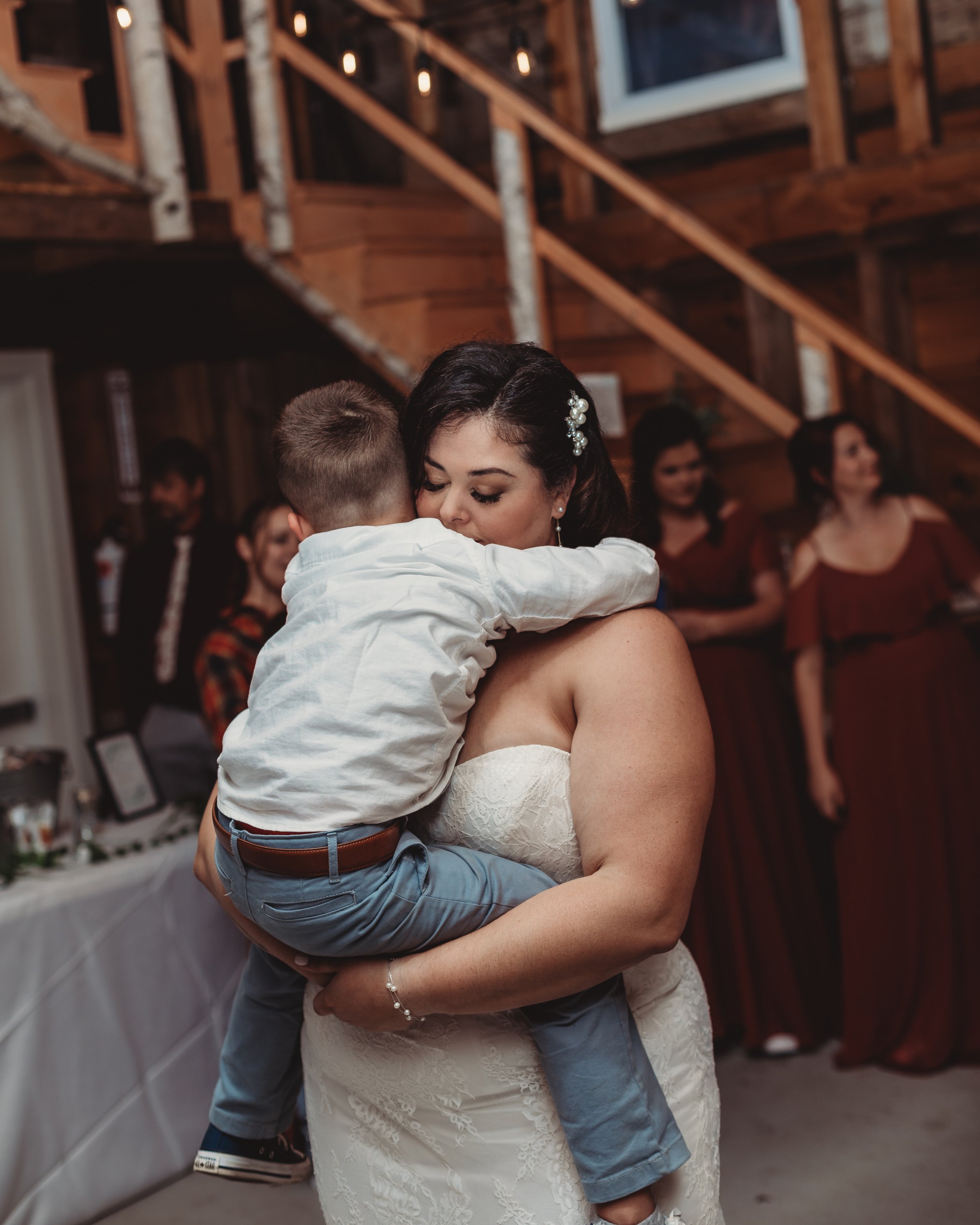 A woman in a wedding dress hugging a young boy at a celebration in a rustic wooden venue with groups of women in matching dresses in the background.