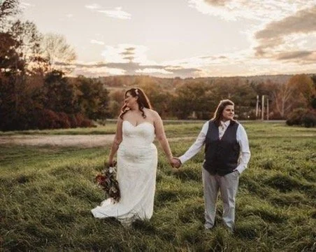 A woman in a white wedding dress holding a bouquet walking hand-in-hand with a man in a vest and dress shirt in a grassy field during sunset.