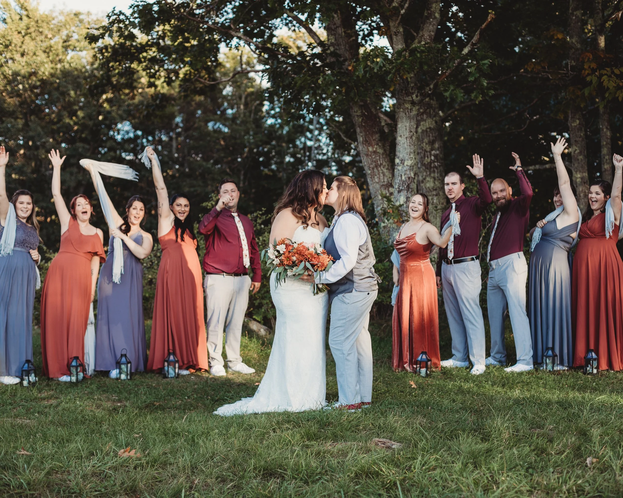 A same-sex couple kissing during their outdoor wedding ceremony, with friends celebrating in the background on a grassy area with trees.
