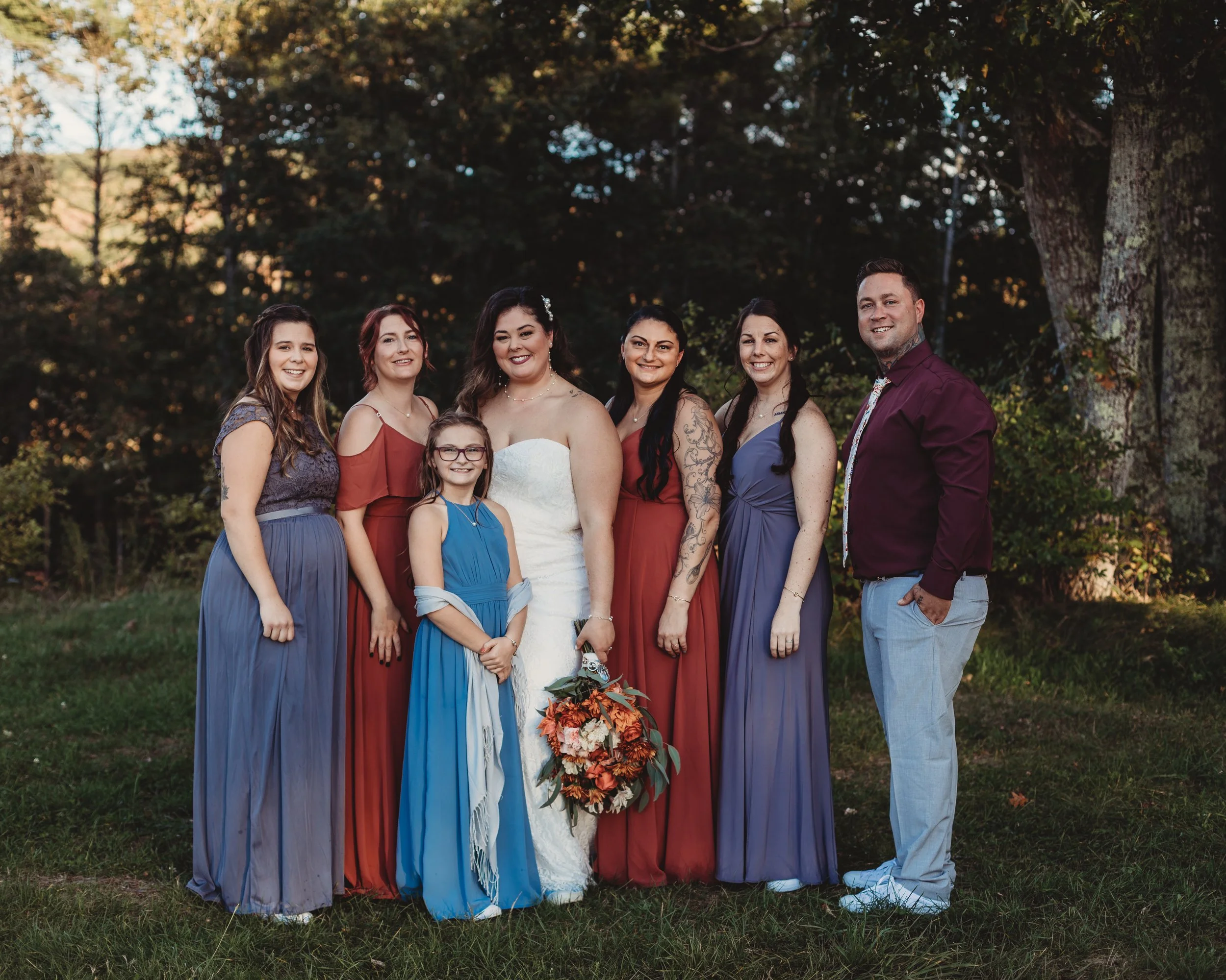 A group of seven women and one man standing outdoors on grass, posing for a photo, with trees in the background, during the daytime. The women are in colorful dresses, and the man is in a maroon shirt and light gray trousers. The woman in the middle 