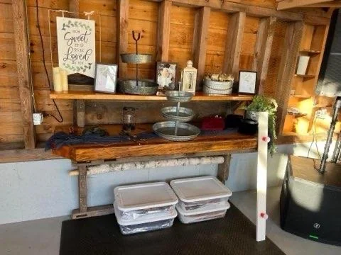 Interior of a rustic wooden shed with shelves holding decorative items, framed photos, containers, and a sign. There are storage bins underneath a wooden table.