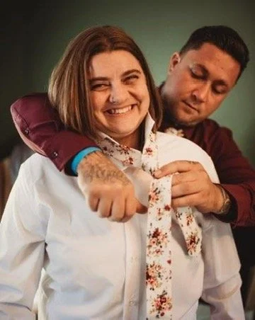 A man playfully puts a tie on a smiling woman, with his arm around her shoulders.