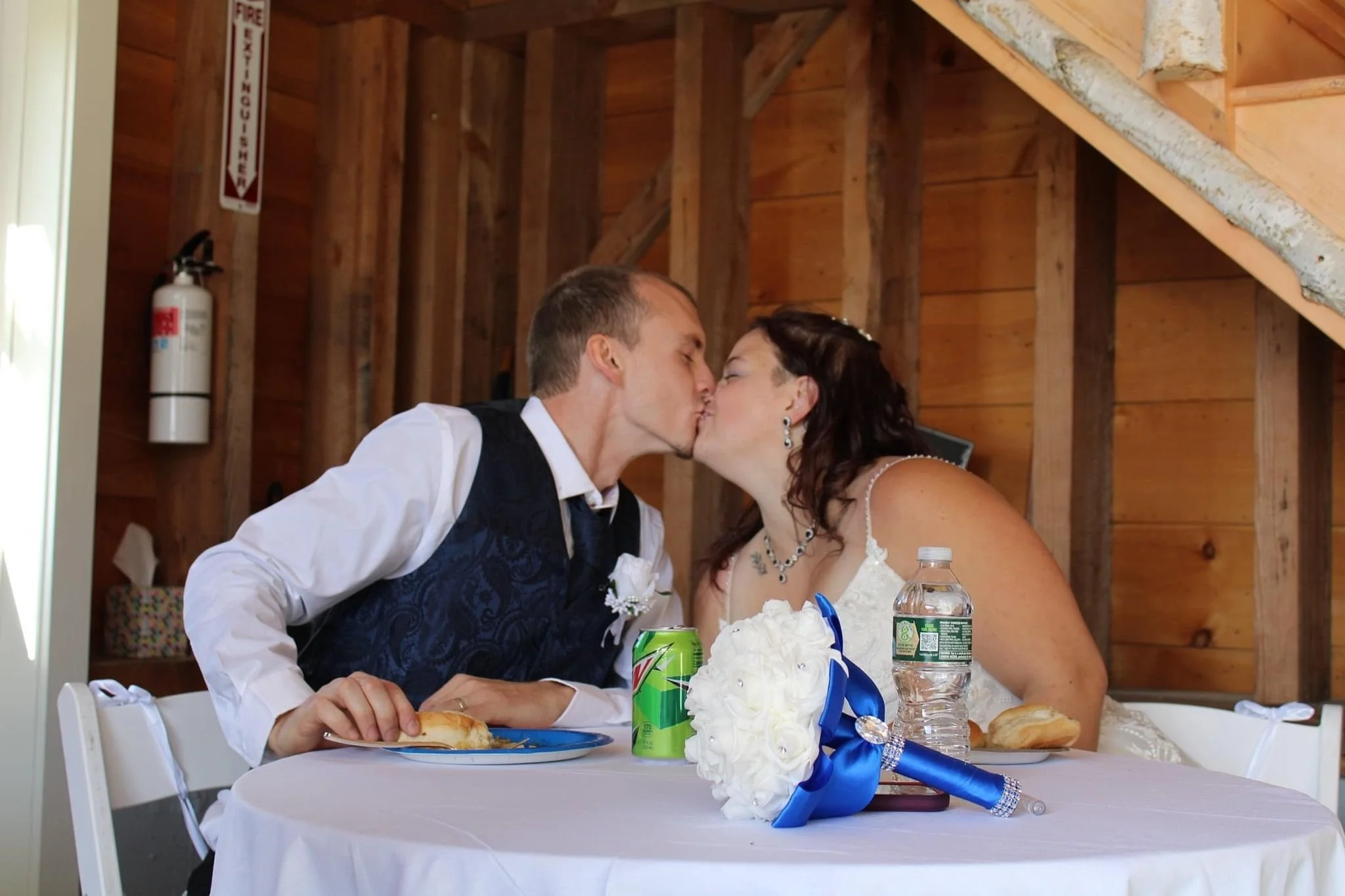 A bride and groom sharing a kiss at their wedding reception, sitting at a table with wedding decor, food, and drinks, inside a wood-paneled venue.
