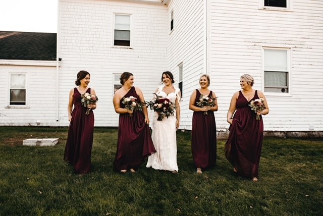 Bride and four bridesmaids standing on a lawn in front of a white house, smiling and holding bouquets, during a wedding celebration.