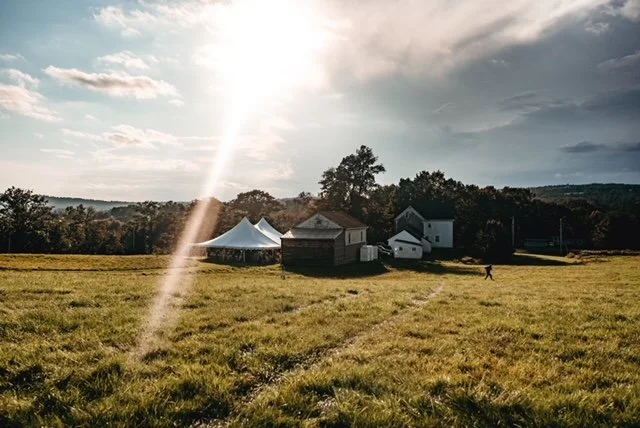 Open field with two white tents, a small shed, trees in the background, and a person walking, under a cloudy sky with sunlight.