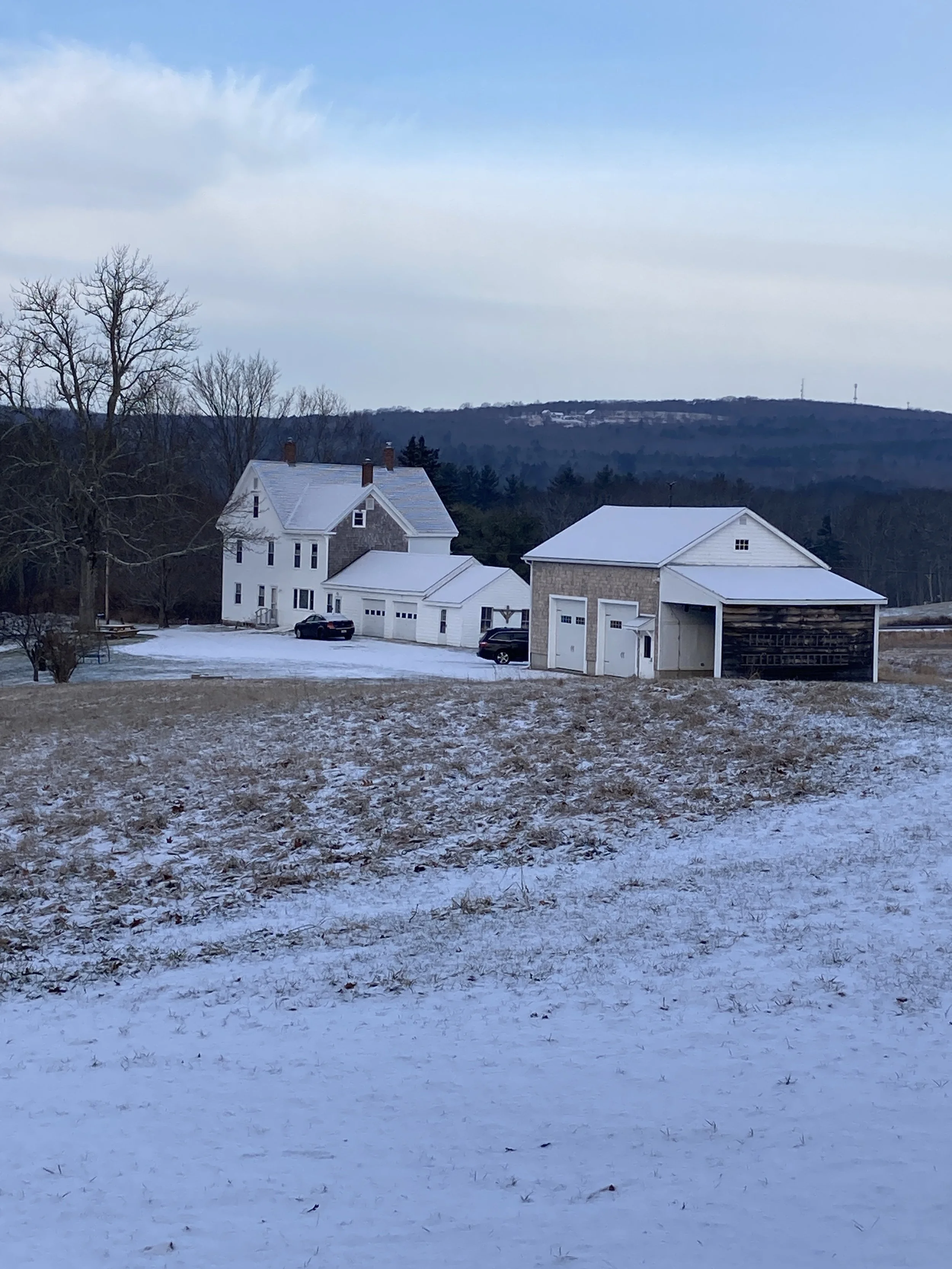 A snow-covered landscape with a large white house, a detached garage, and a barn in a rural area, with hills and trees in the background.