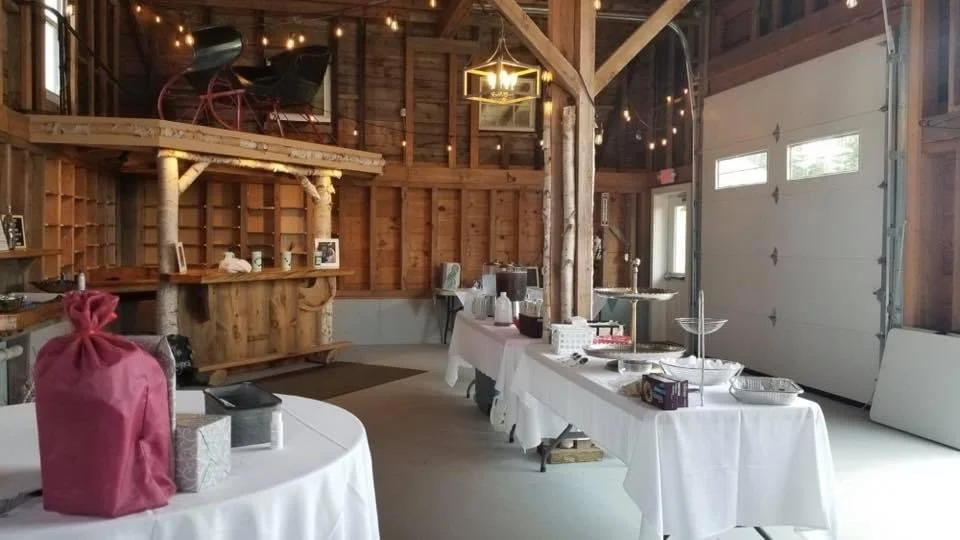Interior of a rustic barn with wooden walls and ceiling, decorated with string lights. Tables are set with white tablecloths for a event, with some food and drink items present. A staircase leads to a loft area with chairs and a small table.