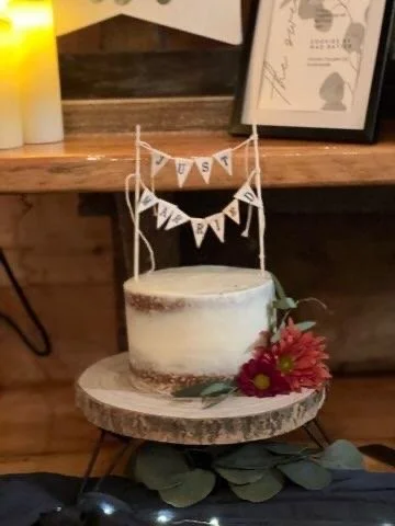Naked birthday cake on a wooden stand with pink flowers and greenery, small decorative banner reading "Just Married" above it, set on a wooden table with candles and framed photos in the background.