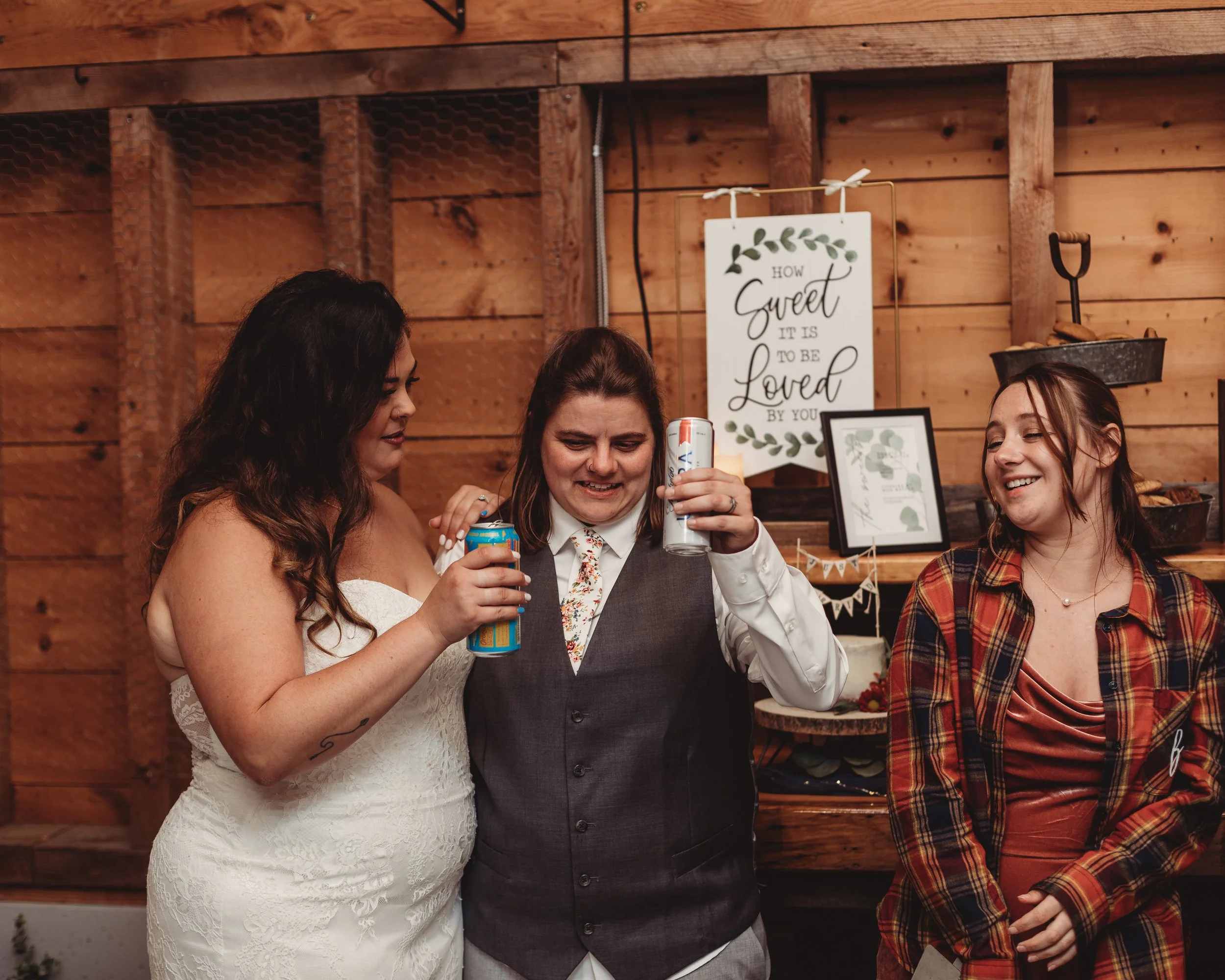 Three women at a celebration, one woman in a wedding dress, holding drinks, standing with two other women, one with a plaid shirt, all smiling in a rustic wooden decorated room.