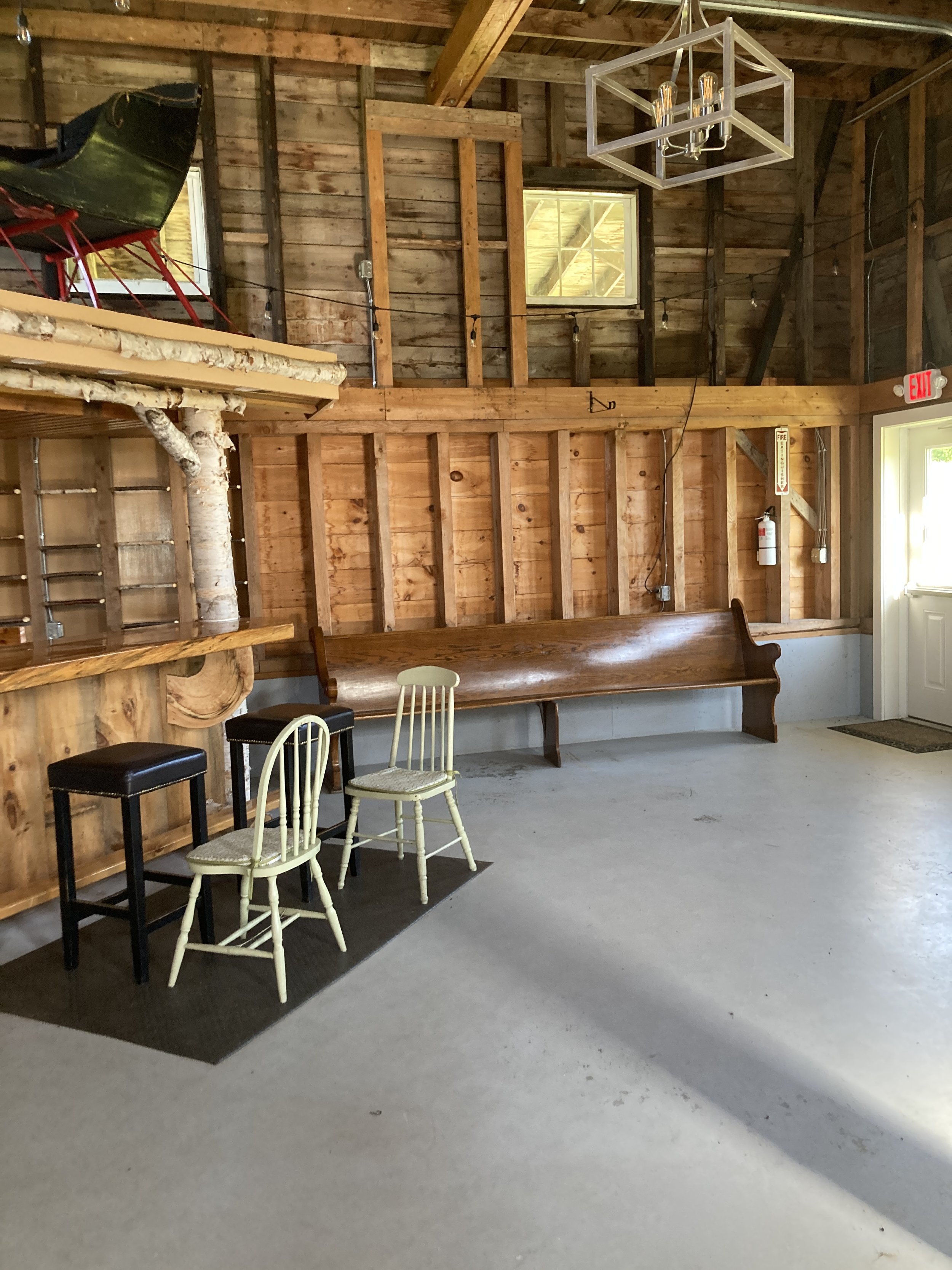 A rustic interior wooden barn with a long wooden bench against the wall, two cream-colored chairs, two black stools, and a black mat on the concrete floor, with a ceiling light fixture and an exit door with an emergency fire extinguisher nearby.