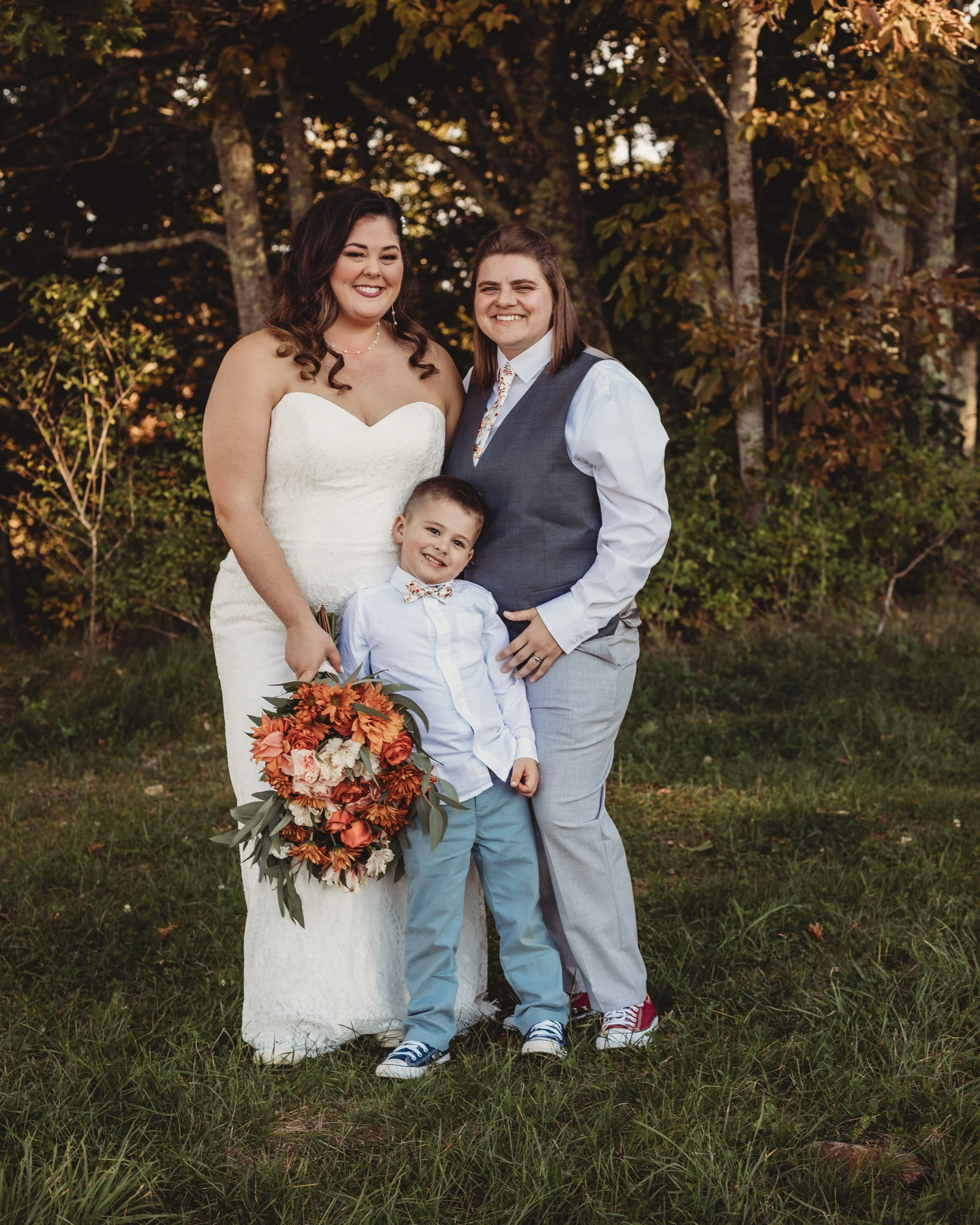 A family of three standing outdoors during fall, with trees and foliage in the background. The woman on the left is in a white wedding dress holding a large bouquet of orange and pink flowers. The woman on the right is dressed in a shirt, vest, and p