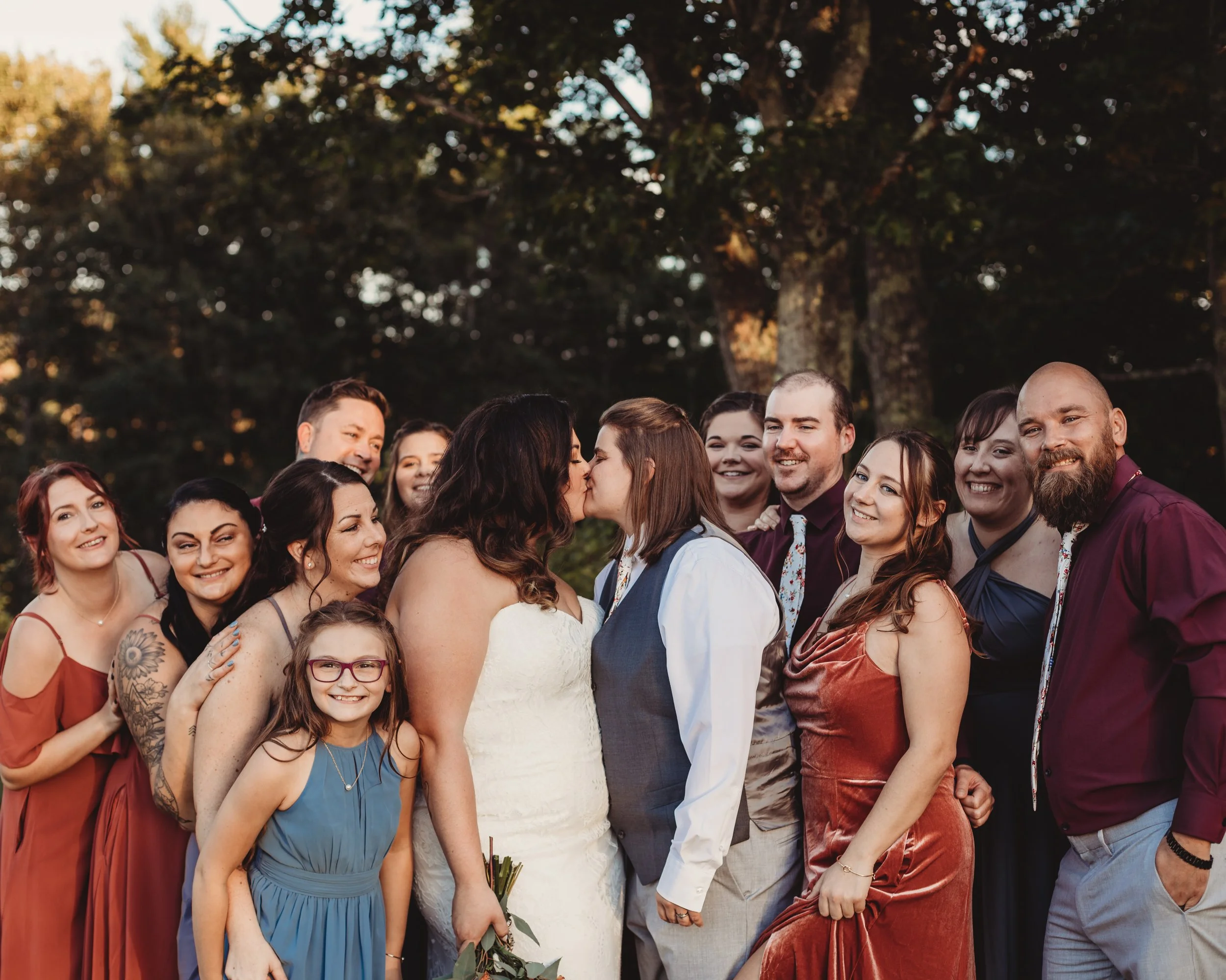 A wedding ceremony outdoors with a group of people, including the bride and groom, exchanging a kiss while surrounded by friends and family during sunset.
