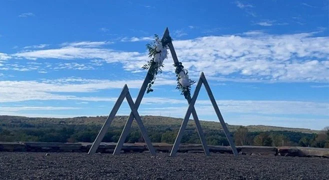 A large outdoor triangular metal structure decorated with white flowers and greenery under a blue sky with scattered clouds.