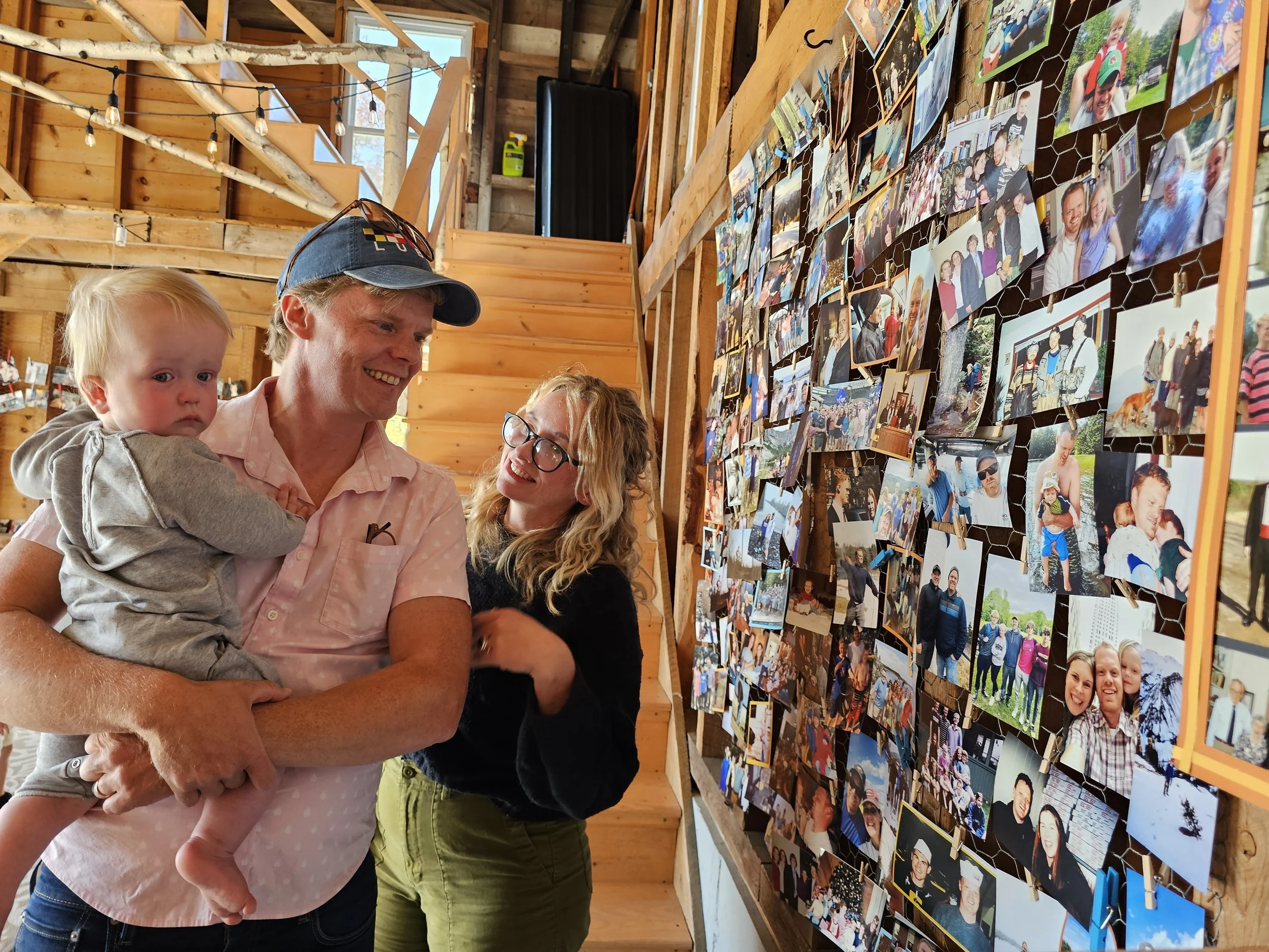 A family of three looking at a display wall covered with numerous photographs of various people, set in a wooden interior with string lights overhead.