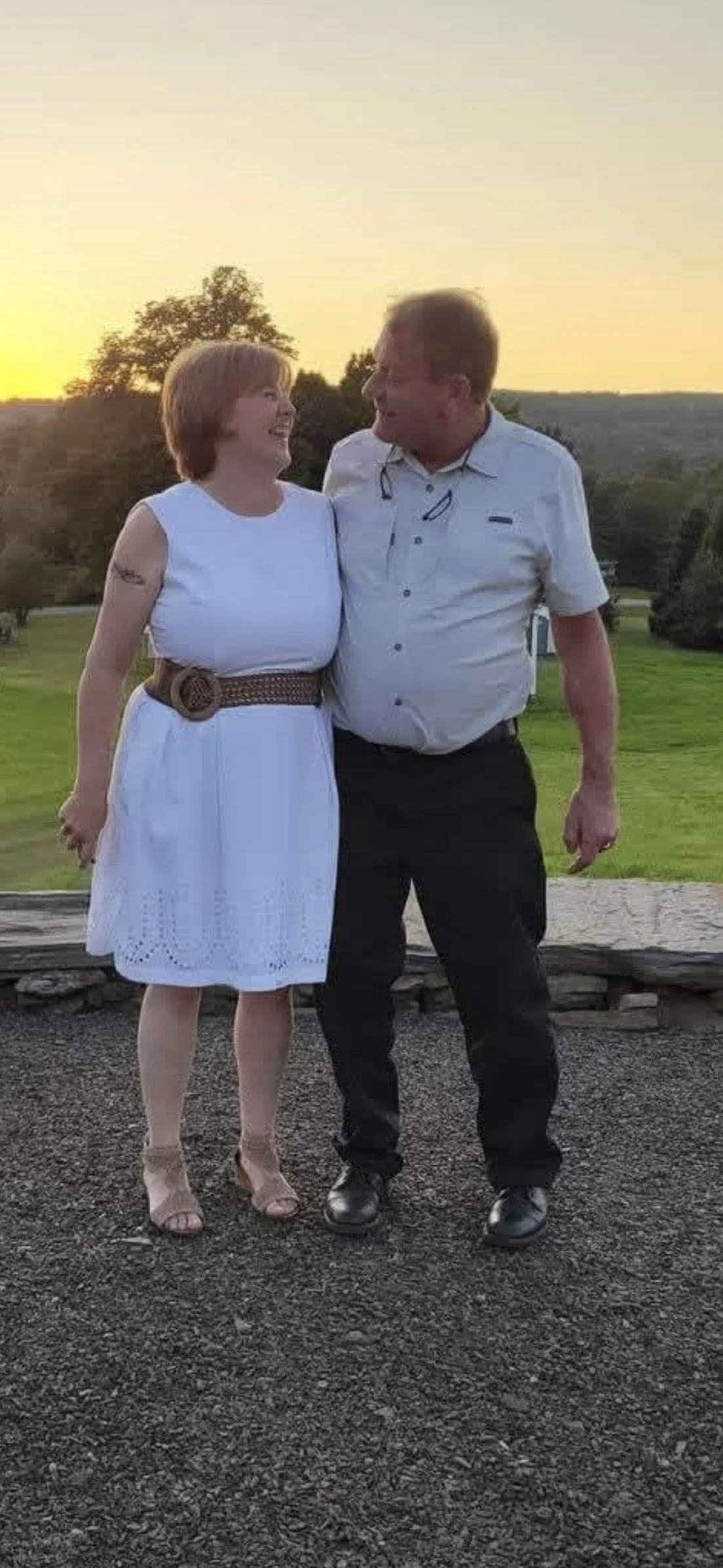 A woman and a man are standing outside during sunset, smiling, with their arms around each other. The woman is wearing a white dress with a brown belt, and the man is wearing a light-colored short-sleeve button-up shirt and dark pants.
