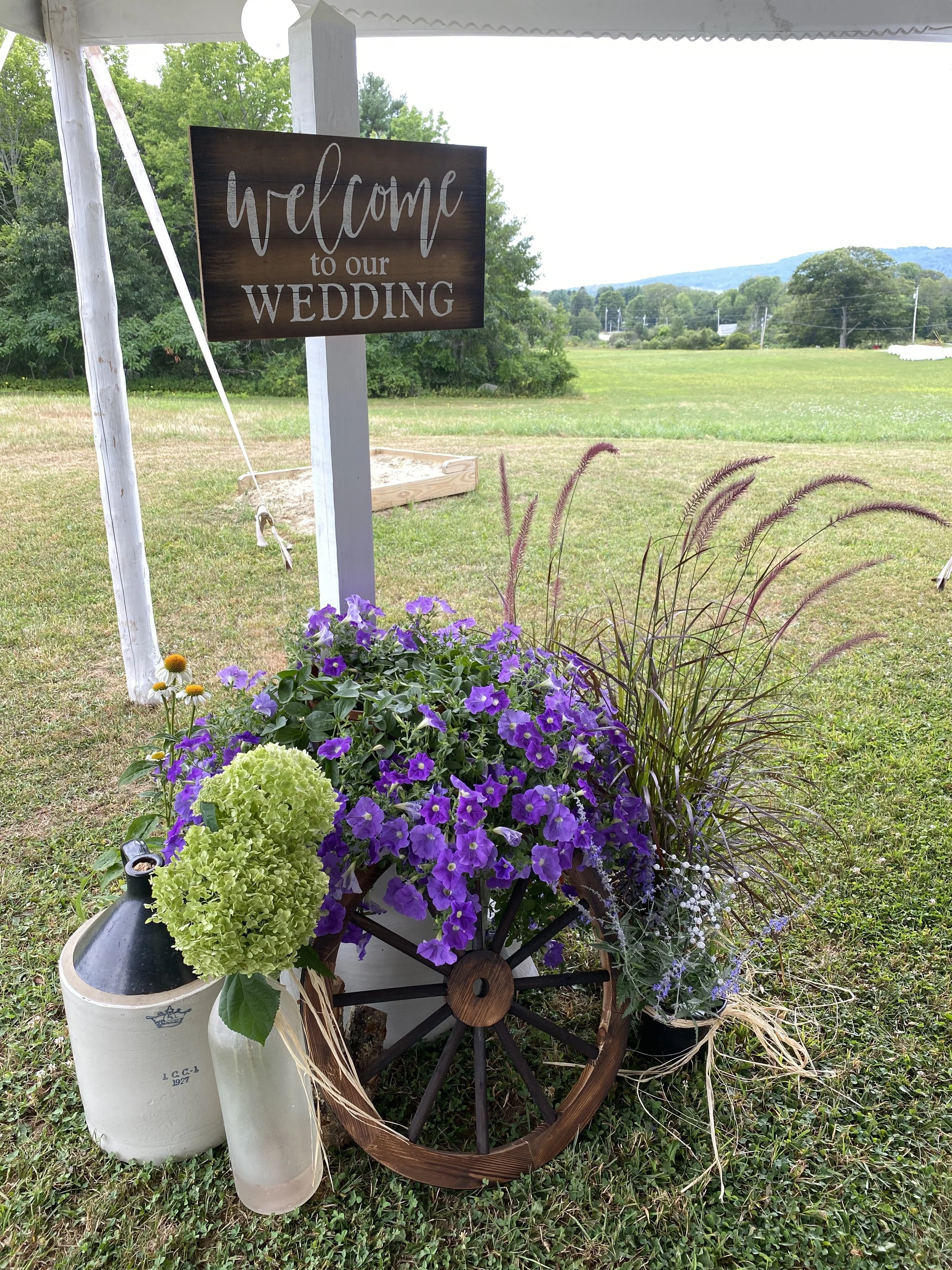 A decorative setup at a wedding with a sign that reads 'Welcome to our wedding,' purple and green flowers in a wagon wheel, a small lantern, and a small bottle, set on a grassy area with trees and open field in the background.