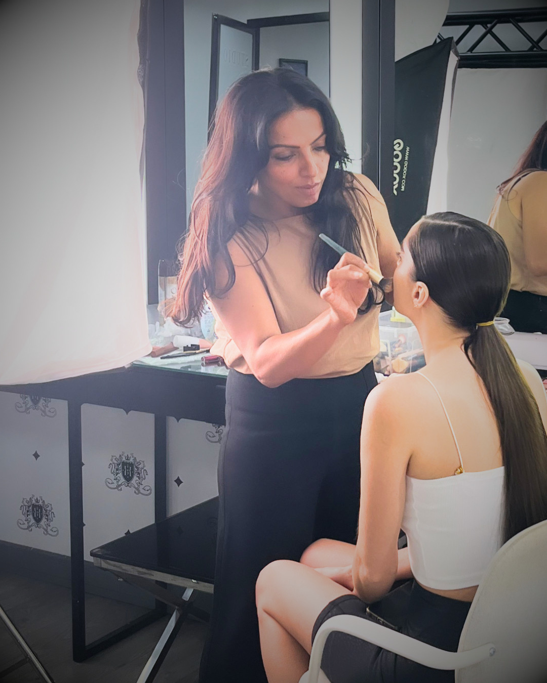 A woman is applying makeup to a seated woman in a salon or studio setting, with professional lighting and makeup equipment in the background.