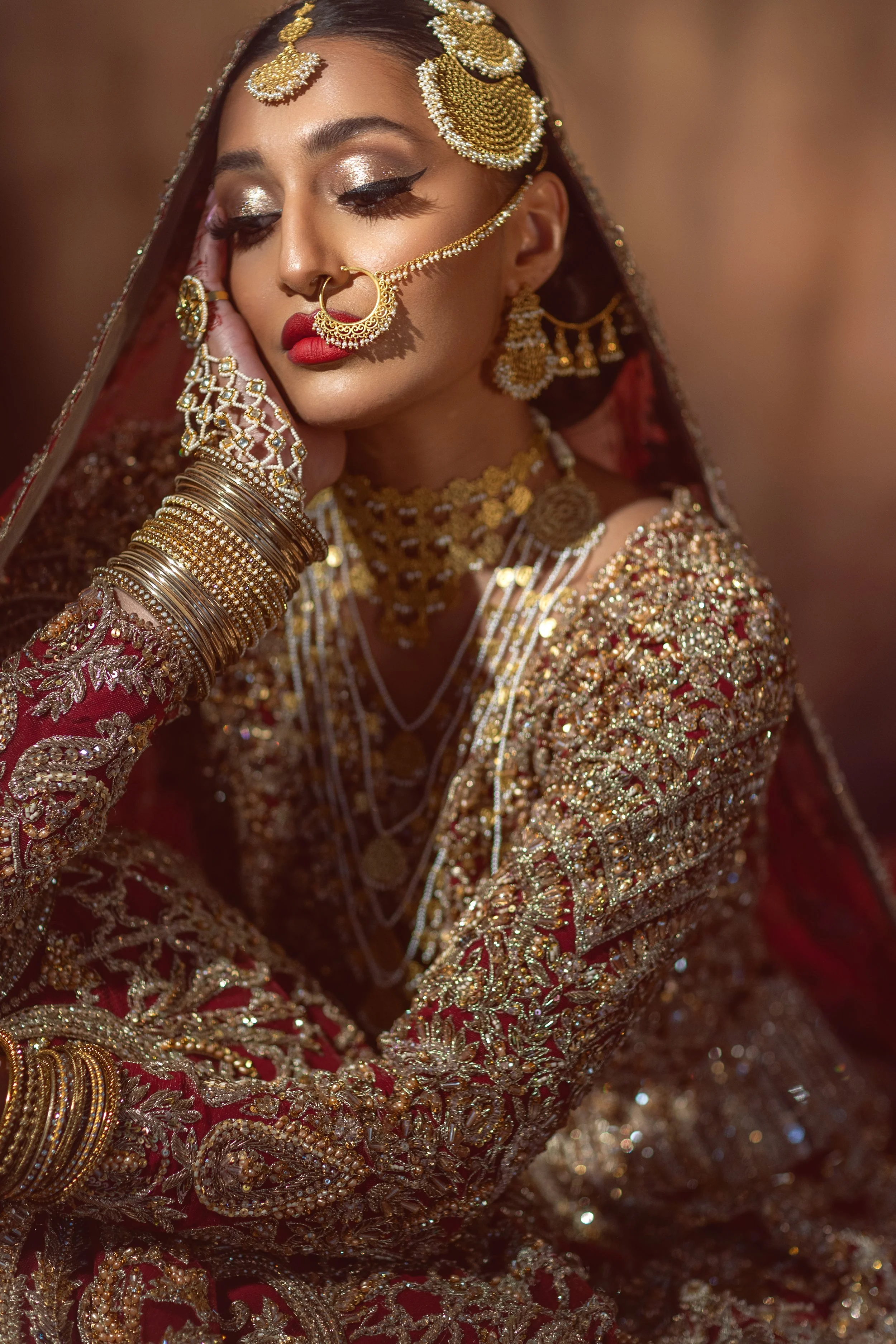 A woman wearing traditional Indian bridal attire with intricate gold and red embroidery, adorned with elaborate gold jewelry including necklaces, earrings, bangles, and a large nose ring, with her eyes closed and one hand resting on her face.