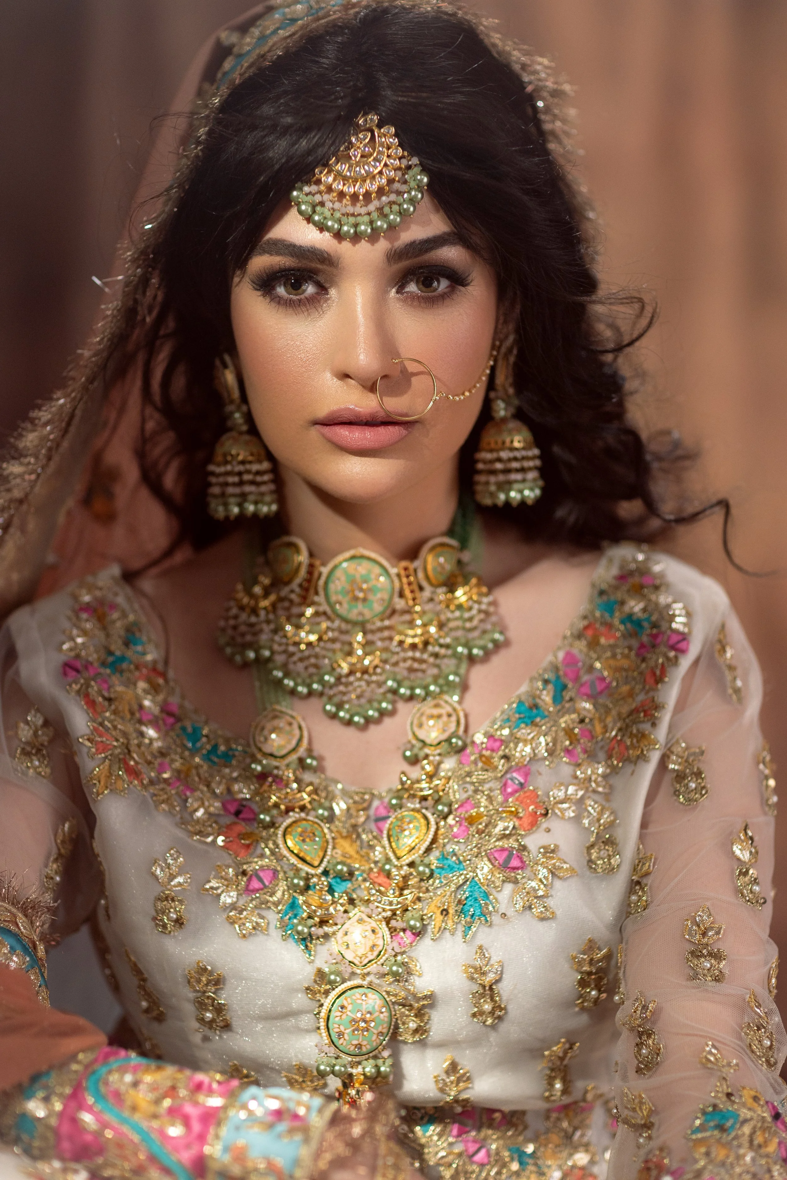 A woman dressed in traditional Indian attire with intricate jewelry, including a maang tikka, nose ring, earrings, and layered necklaces, wearing a white embellished dress with colorful embroidery.